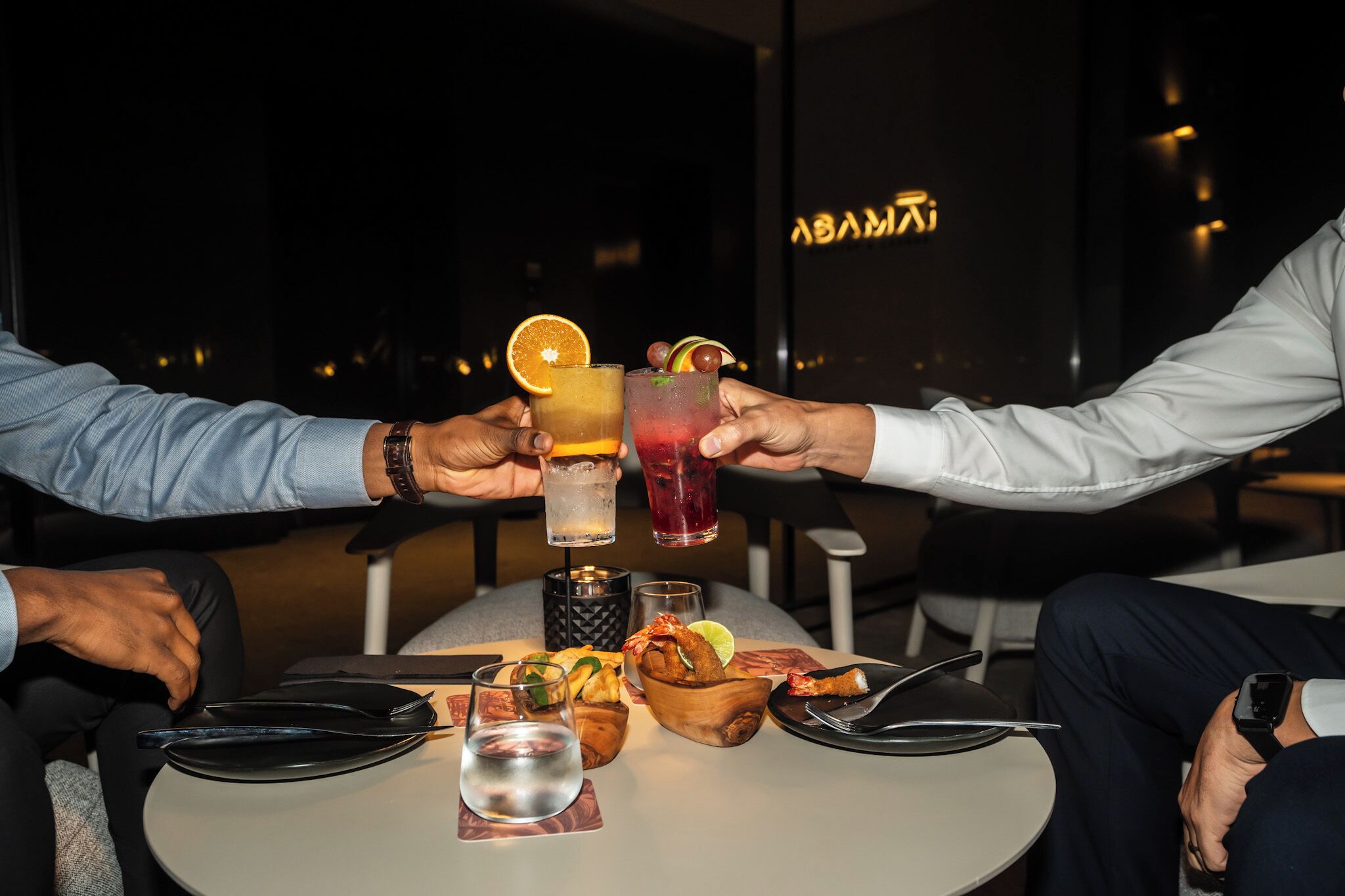 Dos personas brindando con bebidas en una mesa con vista a un paisaje urbano.