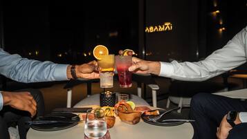 Dos personas brindando con bebidas en una mesa con vista a un paisaje urbano.