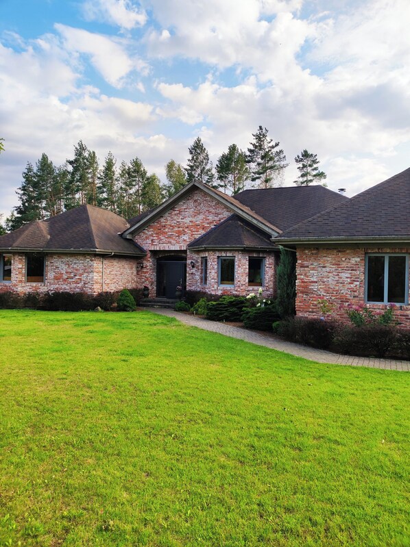 A two-story brick house with a front porch, a covered driveway, and a well-maintained lawn. - Forest apartment in the loop of the river Neris (Verkšionys)