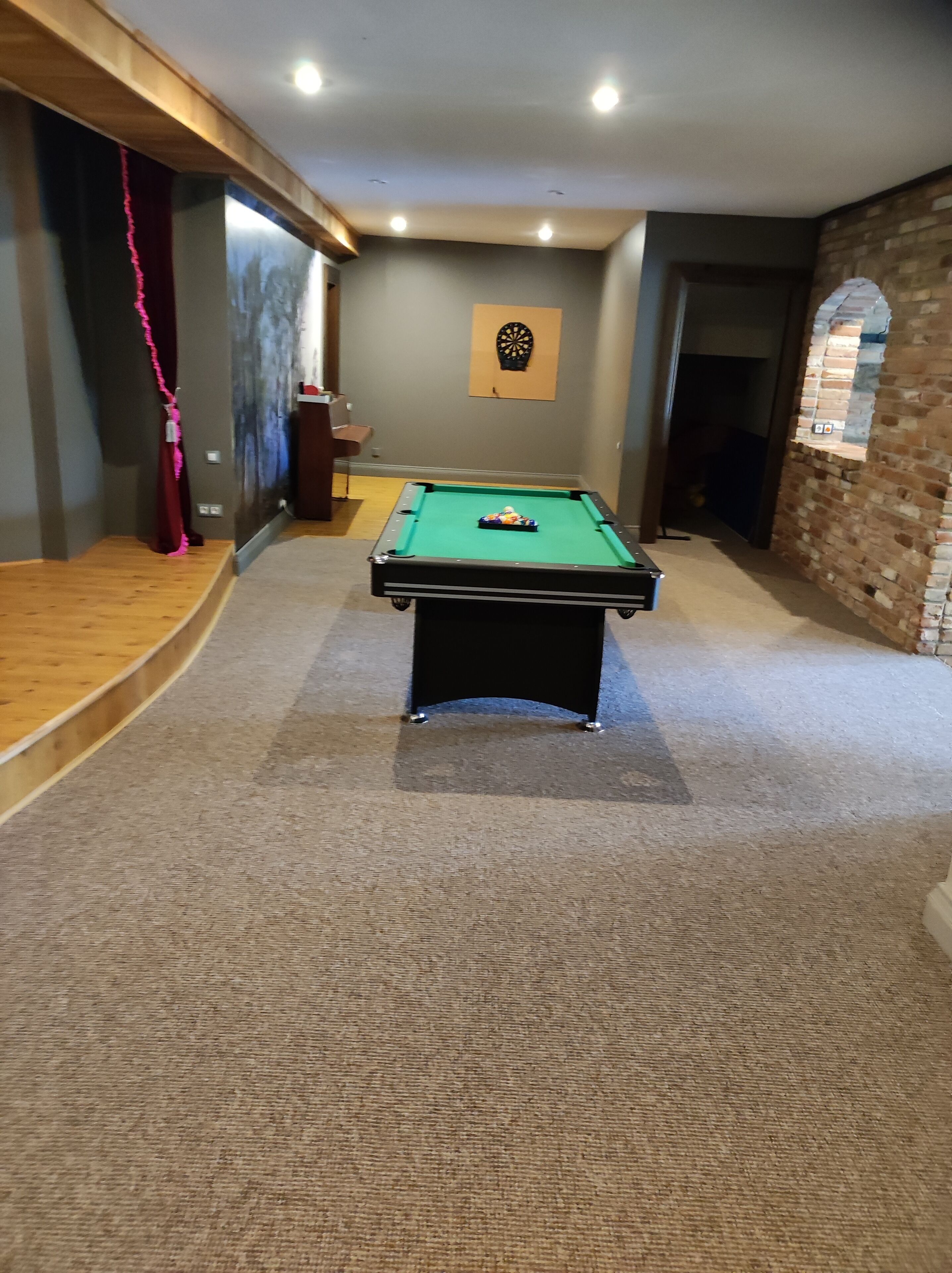 A pool table in a room with a carpeted floor, a dartboard on the wall, and a brick wall with a window.