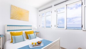 Un dormitorio con cama, una bandeja de desayuno, una ventana con vistas al mar y una obra de arte con motivo de sol en la pared.