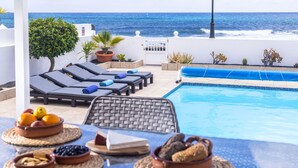 Un área junto a la piscina con sillones de descanso, una mesa con fruteros y vista al océano.