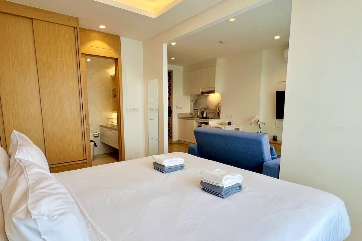A hotel room with a large bed, a wooden wardrobe, a bathroom with a mirror, and a kitchenette in the background.