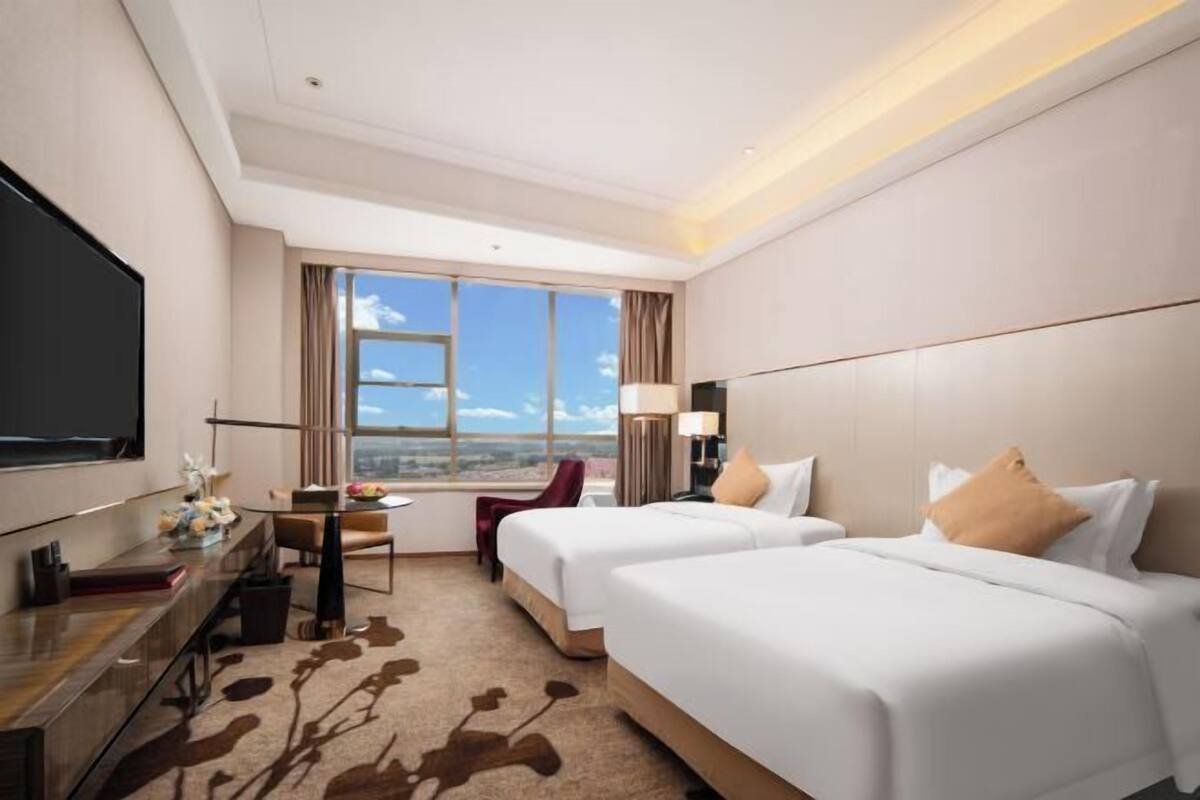 A hotel room with two beds, a desk, a TV, and a large window with a city view.