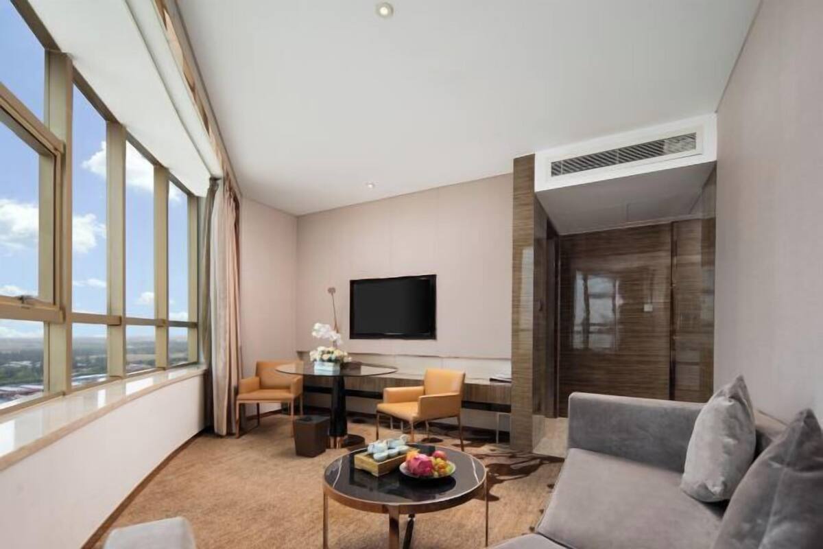 A modern hotel room with a large window, a flat-screen TV, a sofa, a dining table, and a coffee table.