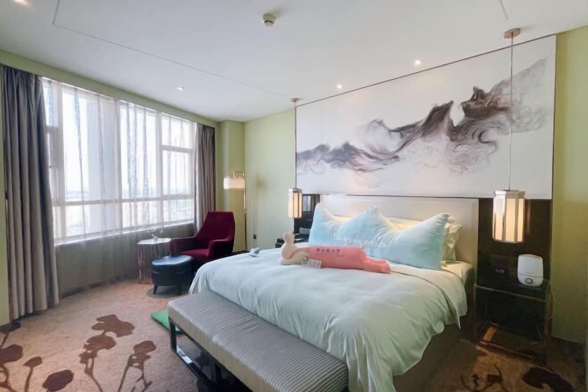 Une chambre d’hôtel avec un grand lit, une banquette, une chaise, un bureau et une grande fenêtre avec des rideaux.