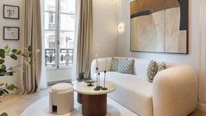 A modern living room with a white sofa, a round coffee table, and a large abstract painting on the wall. - The Lander - Serviced 2BR/2BA - Opéra I (Paris)