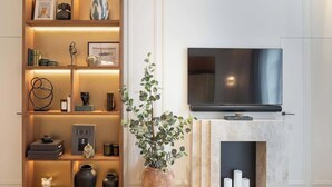 A modern living room with a TV mounted on a marble fireplace, a bookshelf with decorative items, and a large vase with green foliage. - The Lander - Serviced 2BR/2BA - Opéra I (Paris)