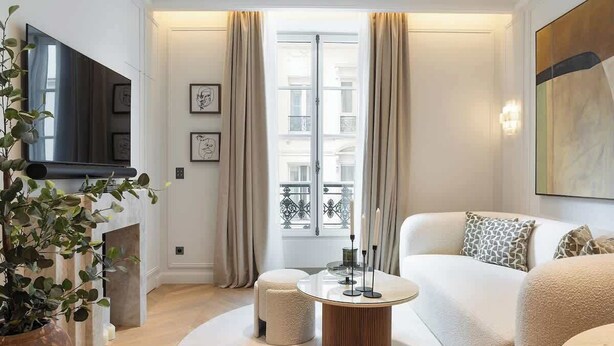 A modern living room with a white sofa, a round coffee table, a TV mounted on the wall, and a large window with curtains. - The Lander - Serviced 2BR/2BA - Opéra I (Paris)