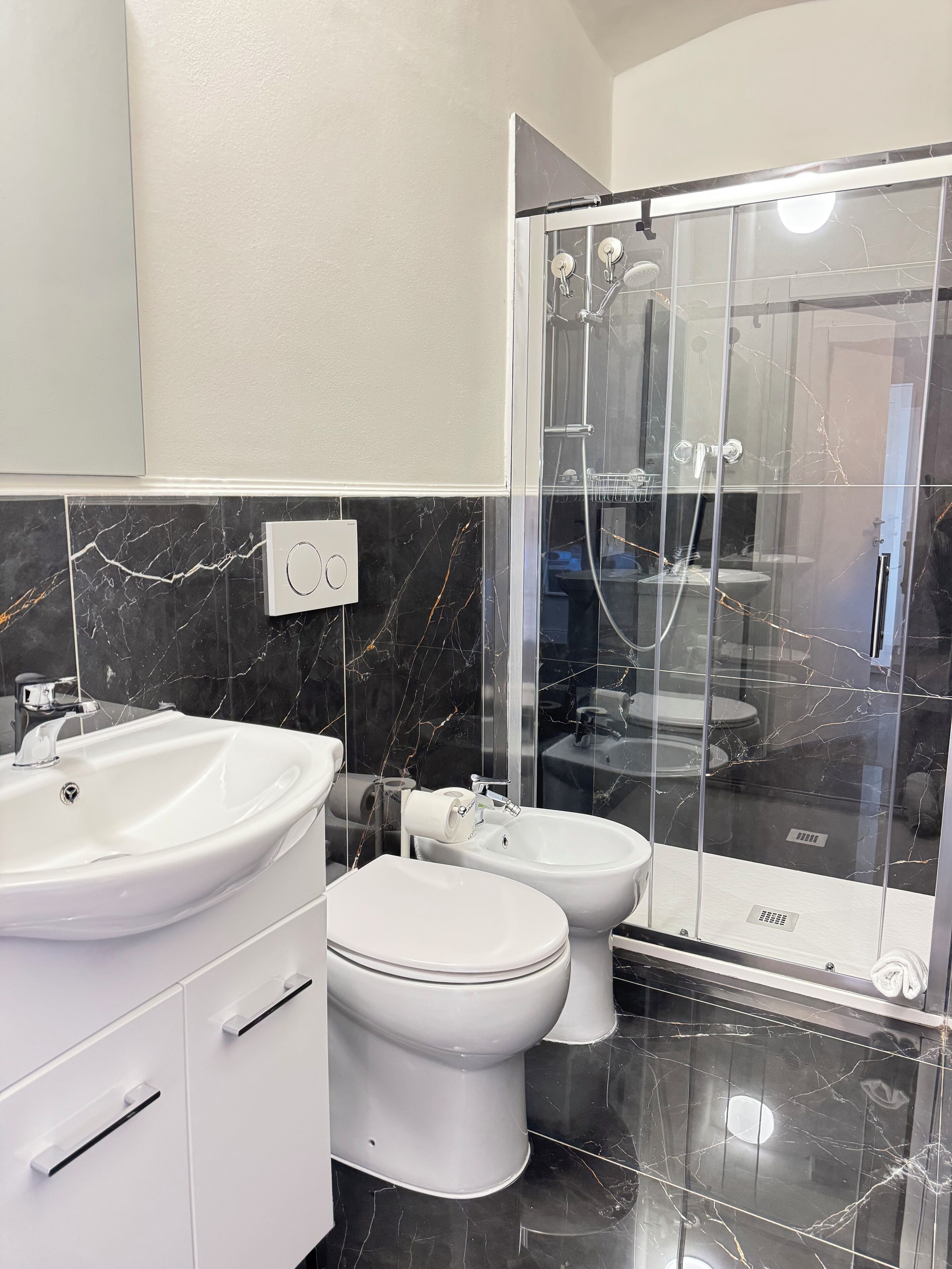 Een moderne badkamer met een wit wastafelmeubel, toilet en bidet, een douche met glazen wanden, en marmeren vloeren.