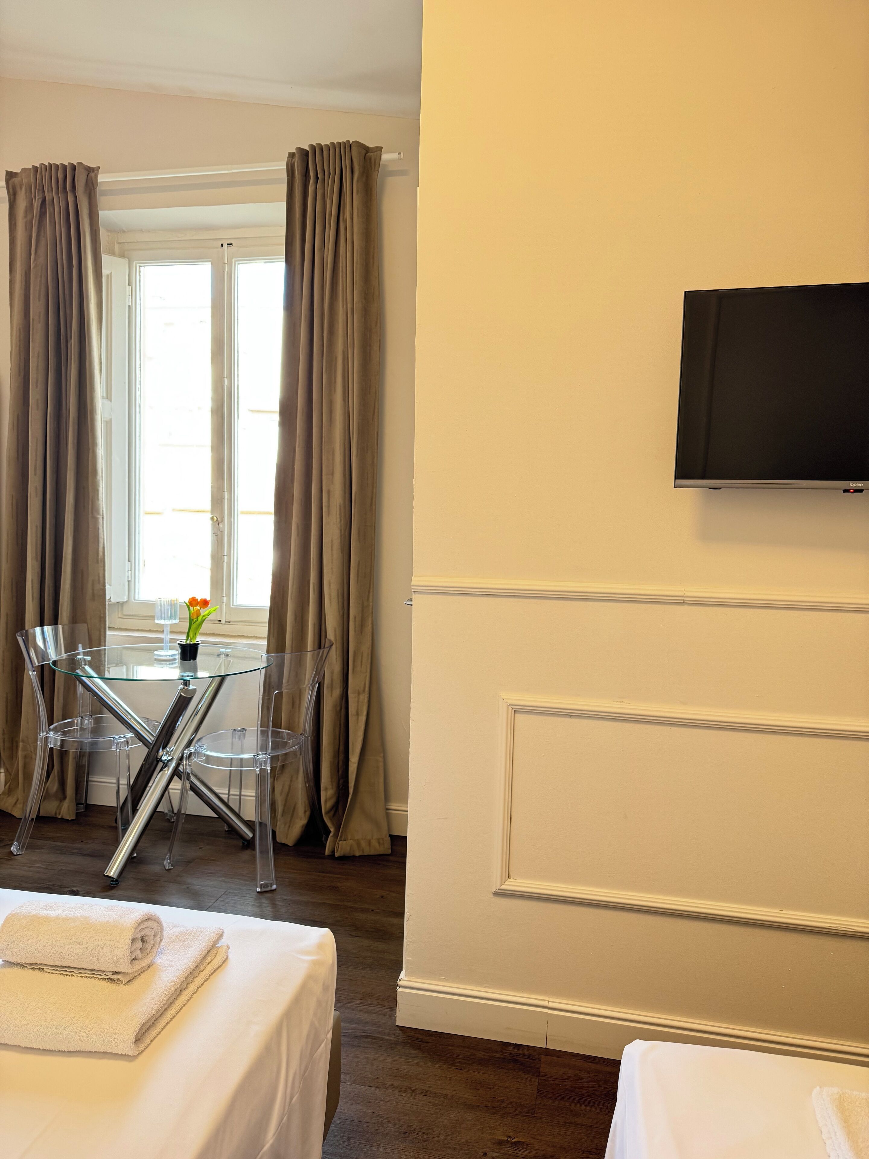 A hotel room with a bed, a glass table with chairs, a television on the wall, and curtains on the windows.