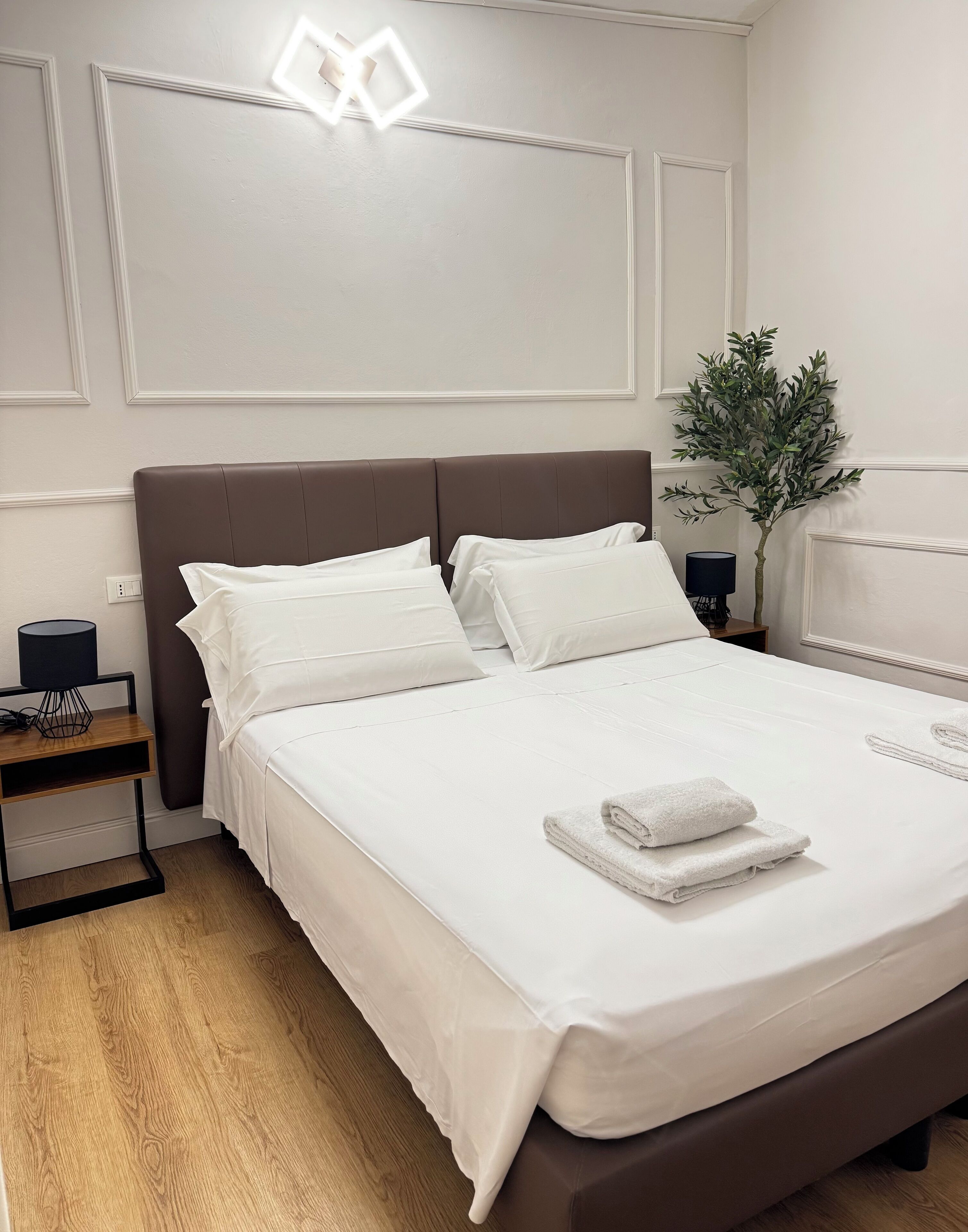 A neatly made bed with white linens and a folded towel, a bedside table with a lamp, and a potted plant on the right.