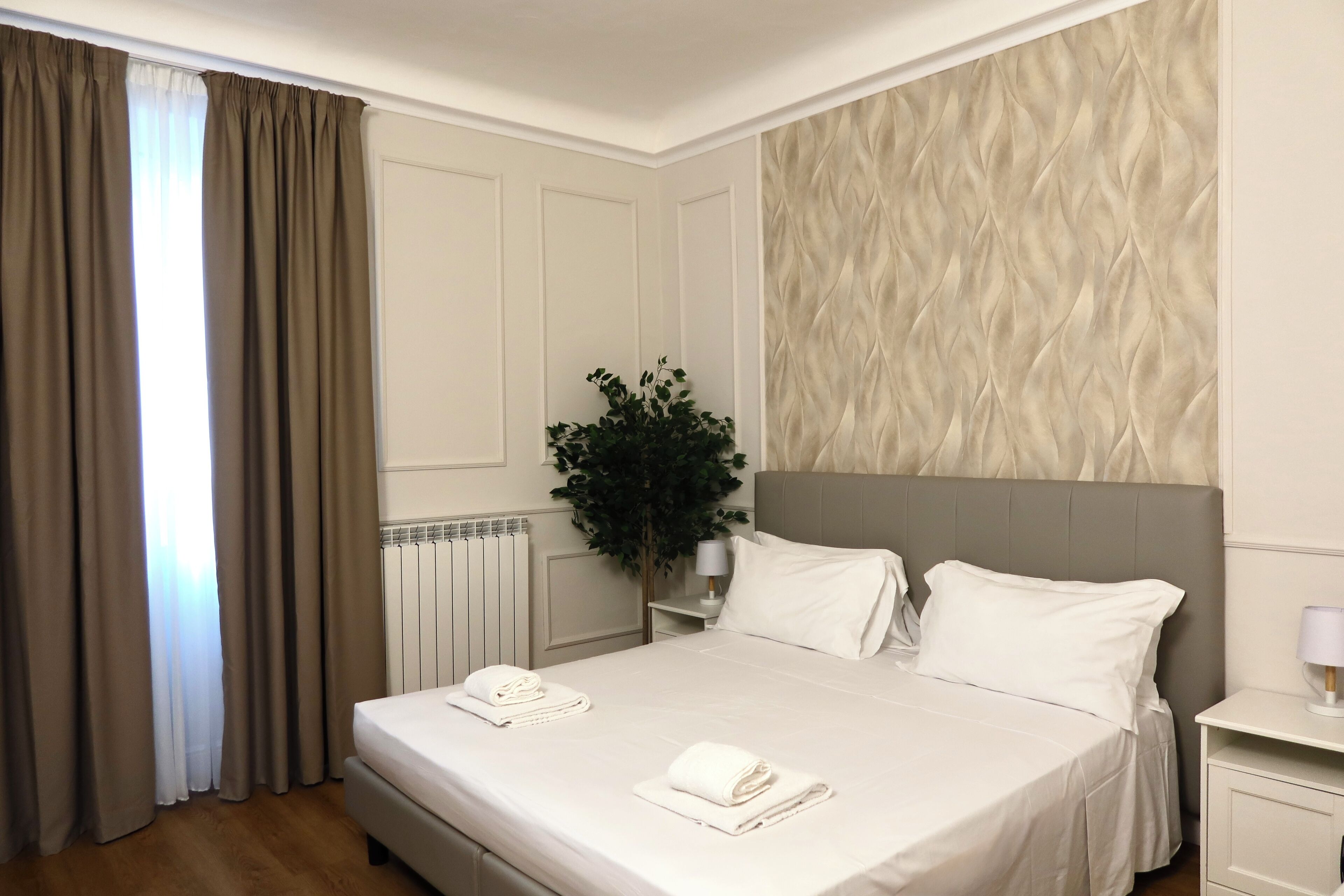 A hotel room with a bed, white linens, a nightstand, a lamp, a potted plant, and a radiator.