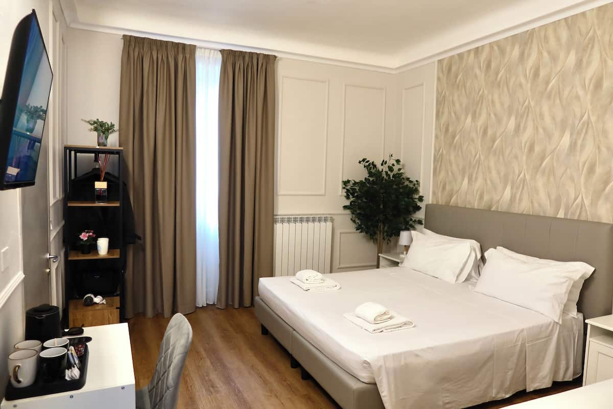 A hotel room with a bed, bedside table, and a desk with a chair. There is a plant on the desk and a radiator on the wall.