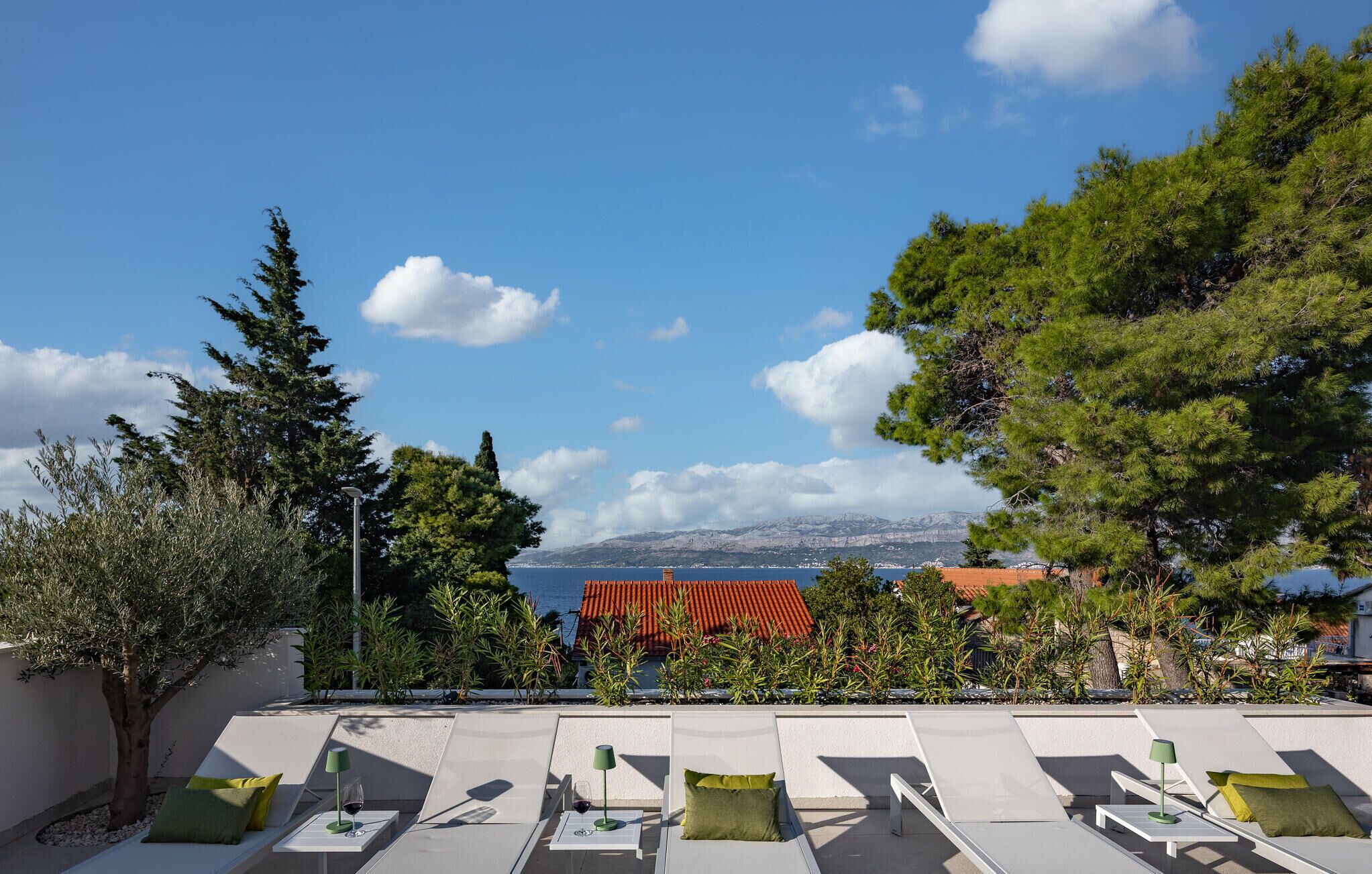 Eine Dachterrasse mit weißen Liegestühlen und grünen Kissen, mit Blick auf die Küste, ein Gebäude mit rotem Dach und Berge in der Ferne.