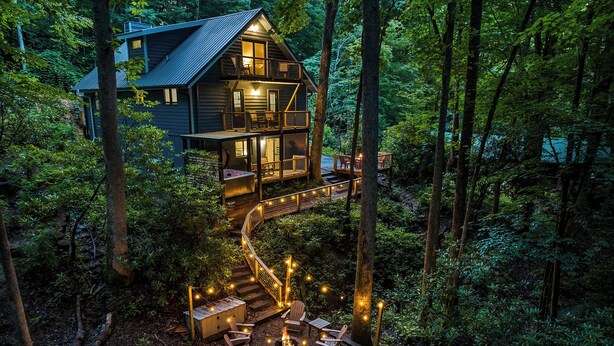 A two-story cabin with a deck and outdoor seating area surrounded by forest.