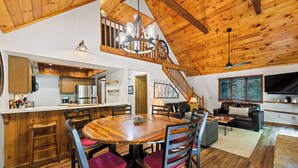 A spacious living area with a wooden ceiling, a dining table with chairs, a staircase leading to an upper level, and a television in the living room.