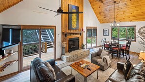 A spacious living area with a fireplace, wooden ceiling, and large windows offering a view of the outdoors.