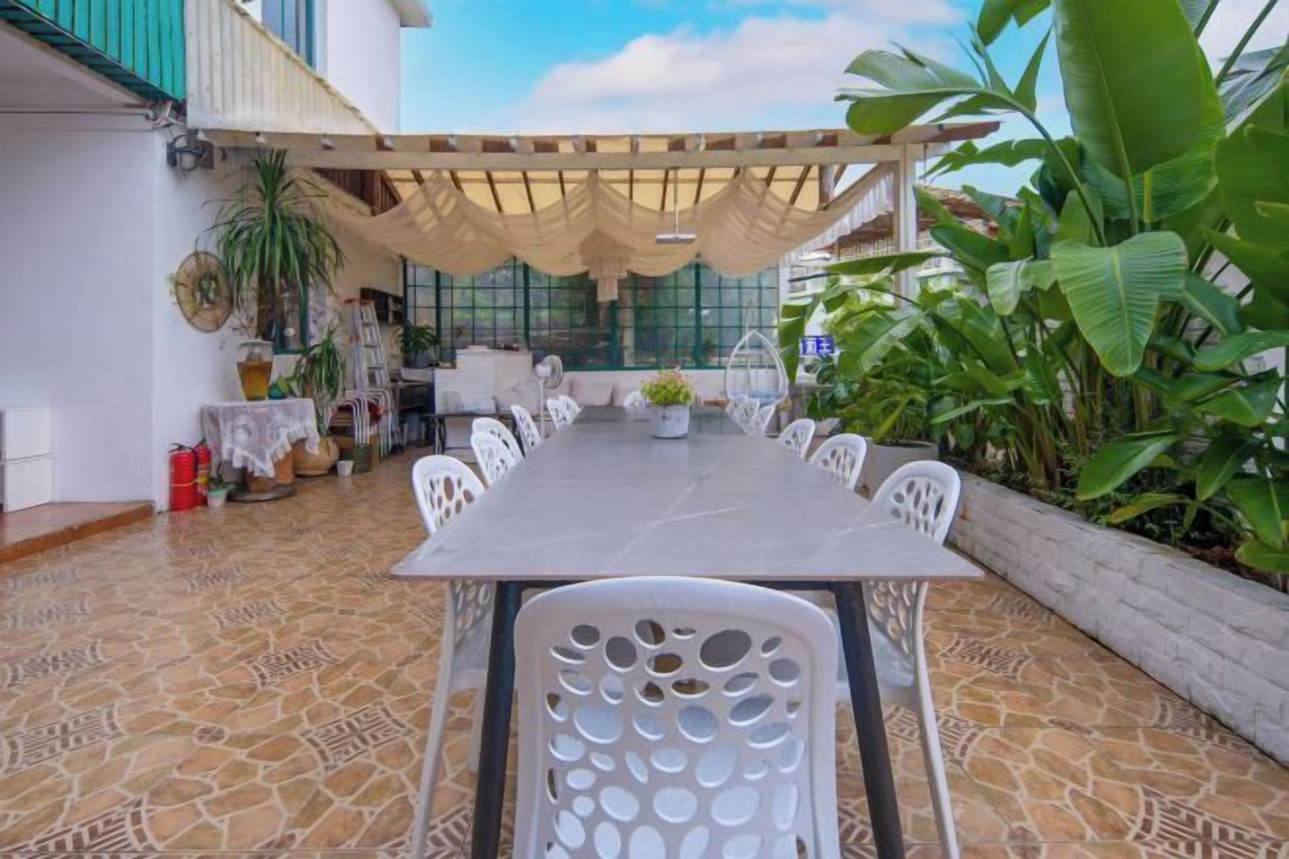 An outdoor dining area with a long table and white chairs, surrounded by potted plants and a pergola.
