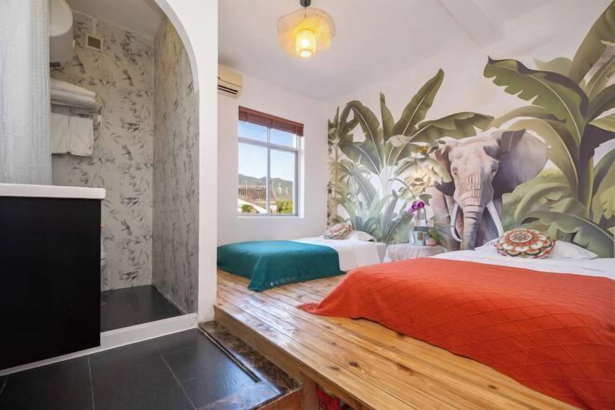 Una habitación con cama, un banco, ducha y un mural de una escena selvática.