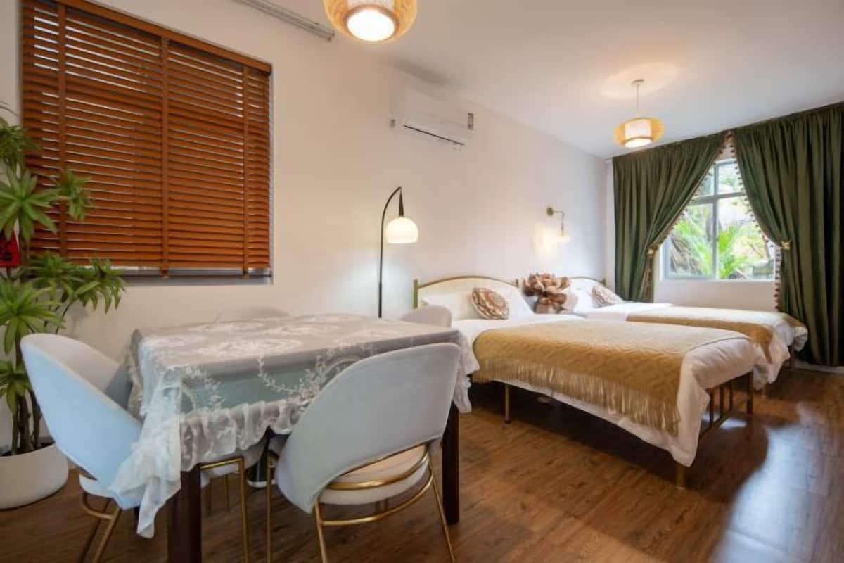 Una habitación con dos camas, una mesa, una silla, una planta en maceta y un ventanal con persianas.