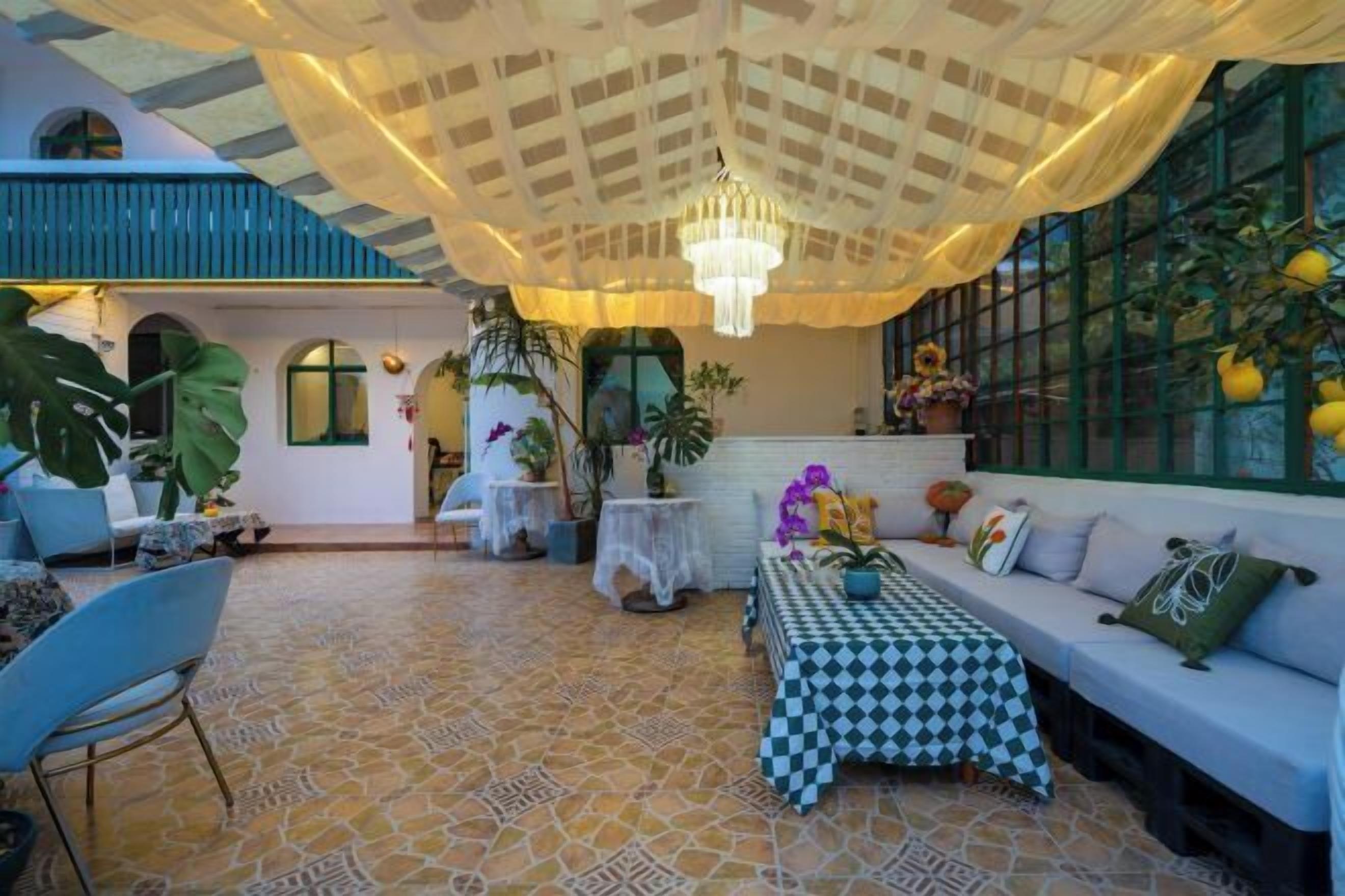 A spacious indoor area with a checkered table, a sofa, and a chandelier. There are potted plants and a large window with a view of greenery.