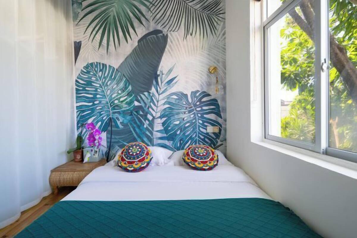 Un dormitorio con un ventanal grande, una cama con colcha verde, dos almohadones decorativos, un mural de hojas tropicales y una pequeña planta en maceta.