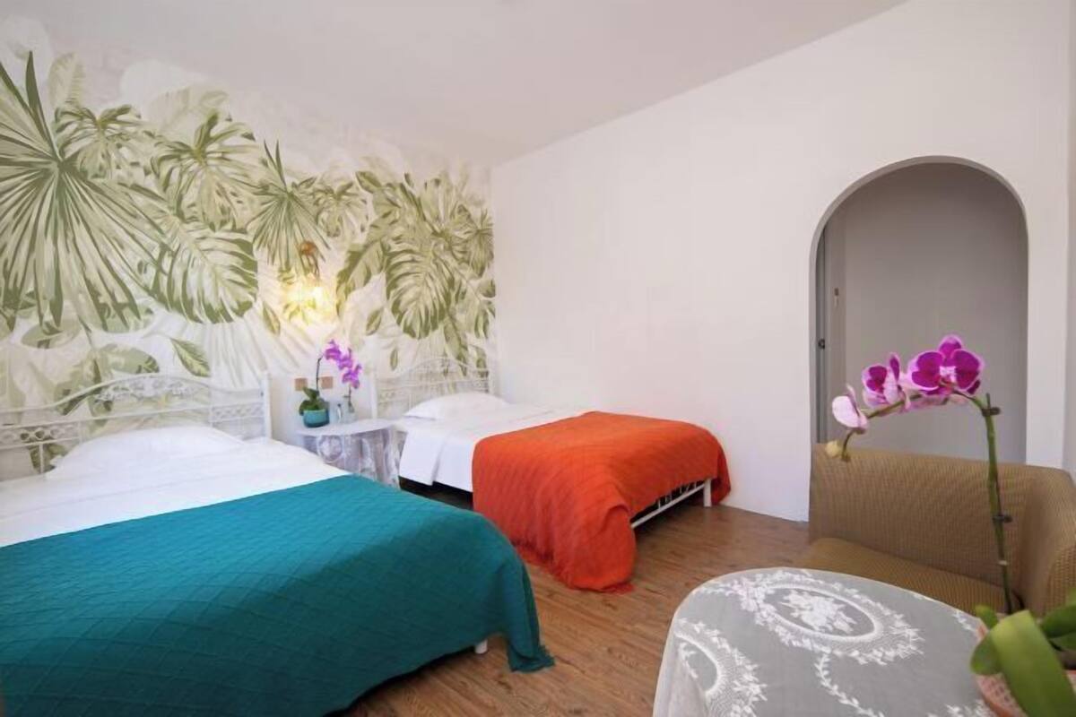 Una habitación con dos camas, una con colcha color turquesa y la otra con colcha naranja. Hay una mesita con un jarrón de flores, una silla y un mural decorativo en la pared.