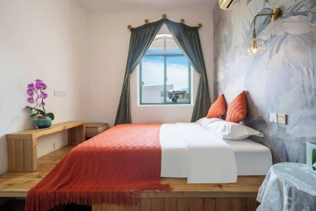 Un dormitorio con cama, ventana con cortinas, lámpara de pared y banco de madera.
