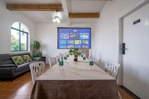 A dining area with a long table, chairs, a projector screen, and a sofa with cushions.