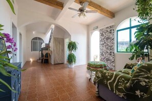 Een ruime woonkamer met een hoge plafond, een plafondventilator, een groot raam met uitzicht, en een bank met bloemenmotief.