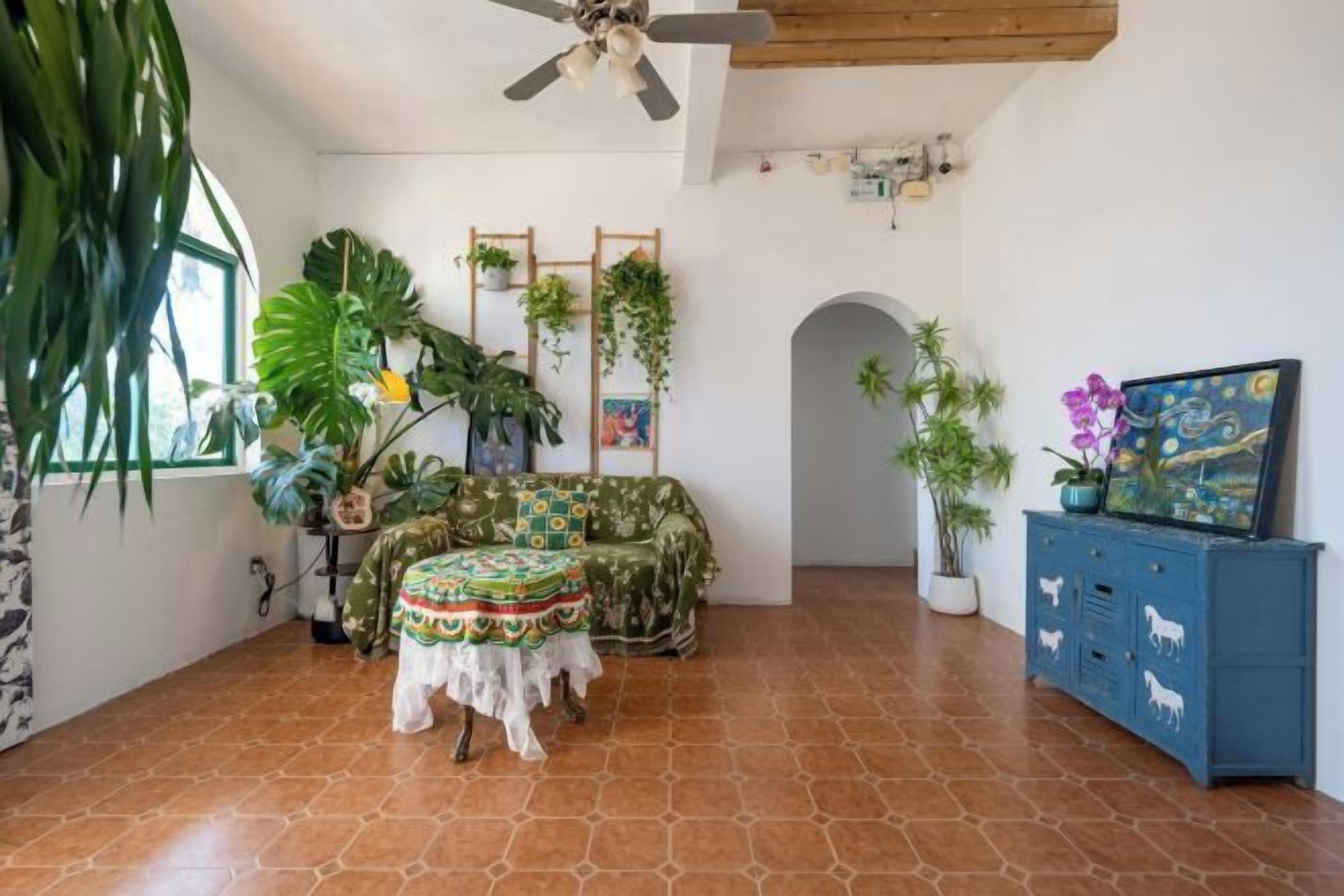 Una habitación con suelo de baldosas, ventilador de techo, sofá, mesa con mantel estampado, un armario azul y varias plantas en macetas.