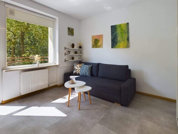 A modern living room with a dark grey sofa, a small white table, and a window with a view of trees.