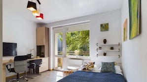 Un dormitorio moderno con una cama grande, un escritorio y una silla. Se ve follaje verde a través de la ventana.