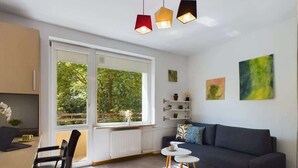 Una sala de estar moderna con un sofá oscuro, una mesa de centro y un comedor con mesa y sillas. Amplias ventanas ofrecen vistas a zonas verdes.