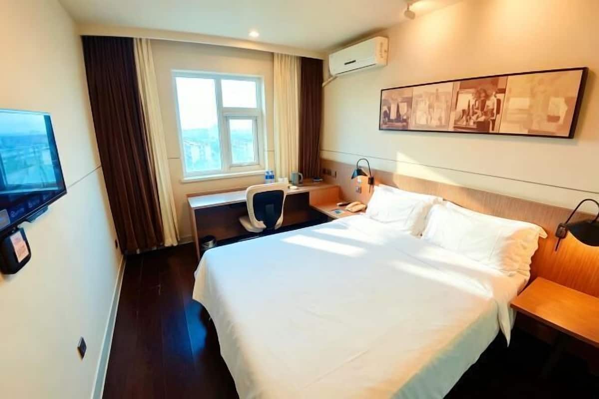 A hotel room with a bed, a desk, a chair, a TV, a window, and a wall-mounted air conditioner.