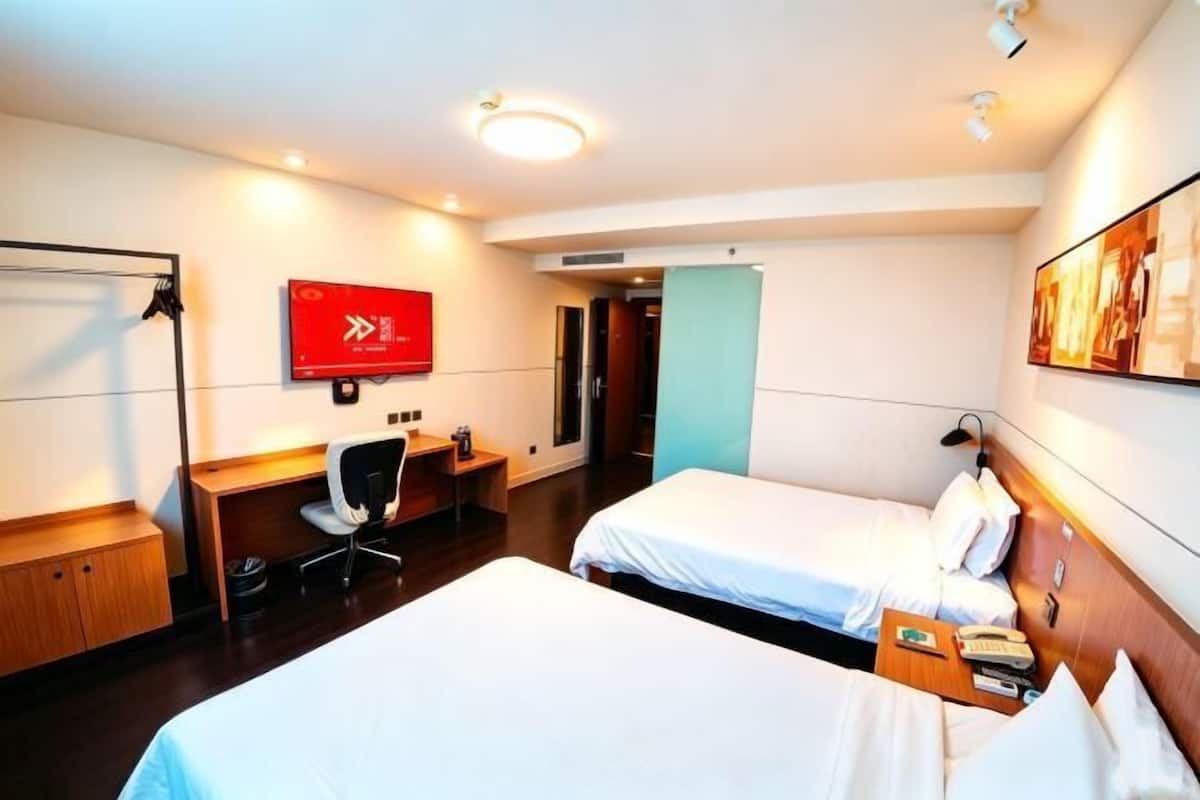 A hotel room with two beds, a desk, a chair, a red emergency exit sign, and a door.