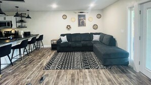 A modern living room with a dark sofa, a patterned rug, and a kitchen area with bar stools.