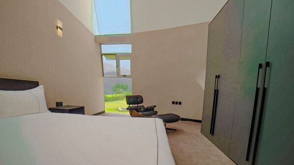 A modern bedroom with a large bed, a desk, a chair, and a wardrobe. There is a window providing a view of greenery outside. - Chebera Elephant Paw Lodge (Konta Koysha)