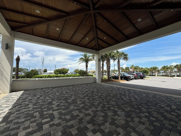 An open-air covered area with a tiled floor, palm trees, and parked cars.