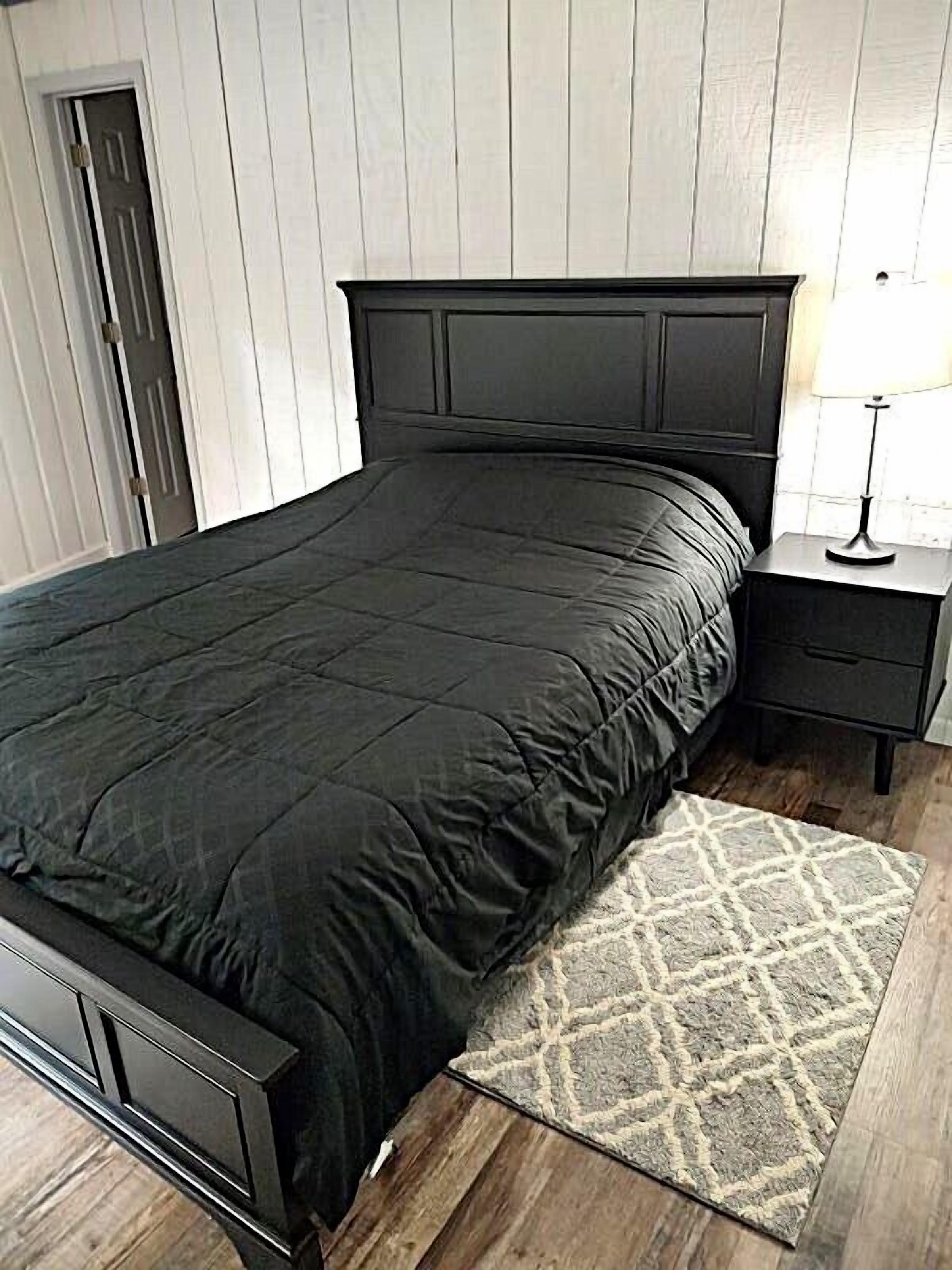 Un dormitorio con una cama de color oscuro, una mesita de noche con lámpara y una alfombra estampada.