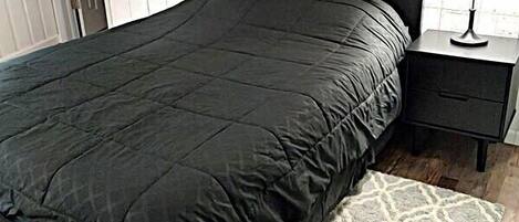 Un dormitorio con una cama de color oscuro, una mesita de noche con lámpara y una alfombra estampada.