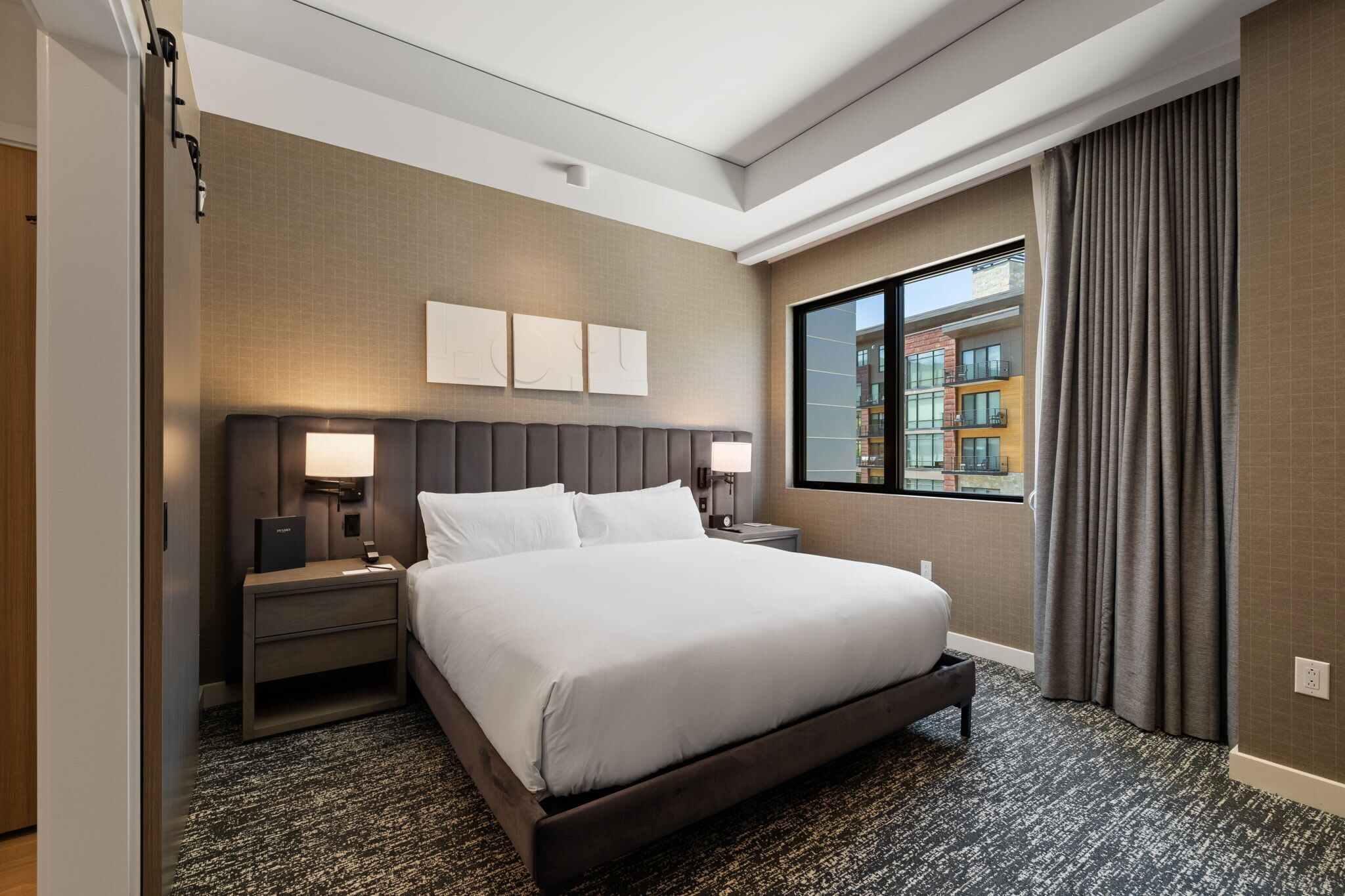 Une chambre d’hôtel avec un grand lit, deux tables de chevet, une fenêtre avec des rideaux et une vue sur les bâtiments environnants.