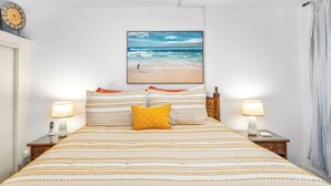 Una cama bien hecha con ropa de cama a rayas y una sola almohada amarilla, flanqueada por mesitas de noche con lámparas y un cuadro colgado en la pared con una escena de playa.