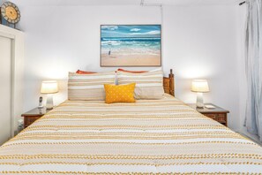 Una cama bien hecha con ropa de cama a rayas y una sola almohada amarilla, flanqueada por mesitas de noche con lámparas y un cuadro colgado en la pared con una escena de playa.