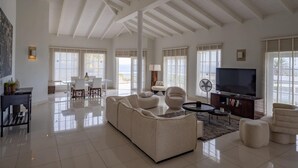 A spacious living room with a high ceiling, large windows, a sofa, a coffee table, a TV, and a bookshelf. - Artiko Luxury Suites - Jan Thiel (Jan Thiel)