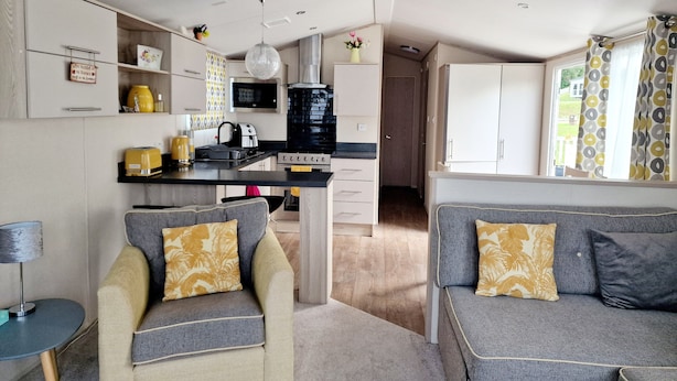 Un espace de vie compact comprenant un coin cuisine, deux canapés avec des coussins à motifs et une fenêtre avec des rideaux.