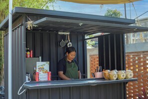 A small outdoor food stall with a metal roof, a person preparing food, and several cooking utensils hanging above. - Bale Taru Pecatu (Pecatu)
