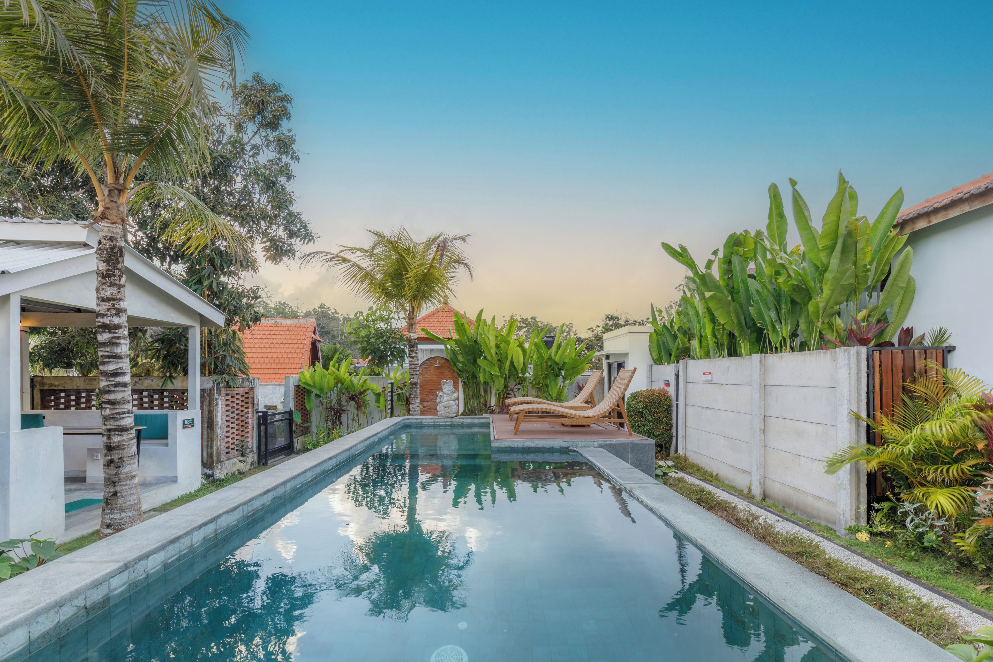 A swimming pool with a clear view of the sky, surrounded by lush greenery and a white building with a red roof.