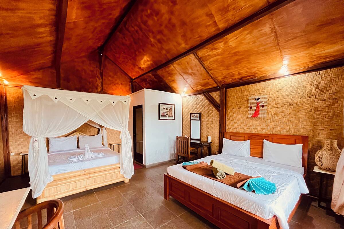 Una habitación con dos camas, una con dosel y otra sin, un banco de madera, una mesita y un espejo en la pared.